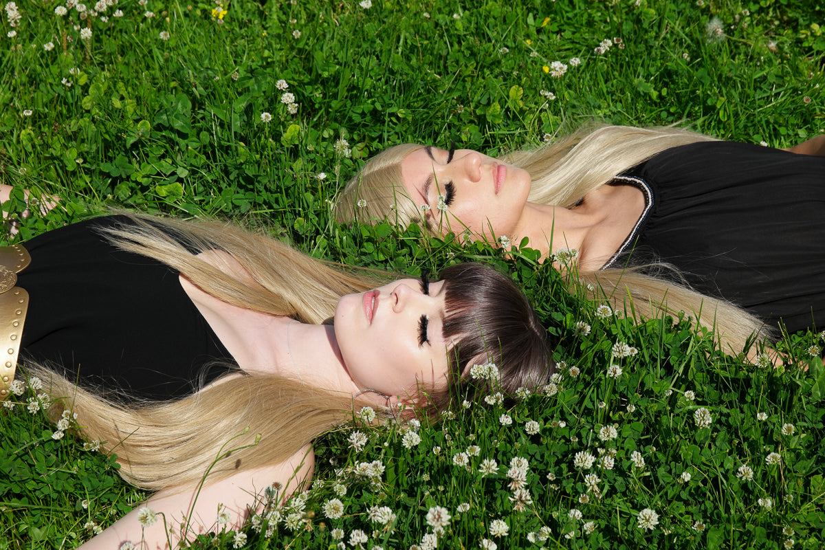 Två kvinnor ligger i gräset med långa blonda peruker som sprider sig över klöverblommorna, den ena med lugg och den andra med mittbena, båda i svart klänning och med ett drömlikt uttryck.